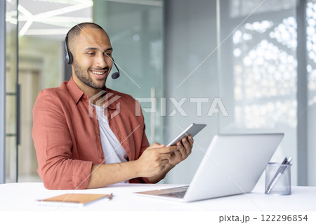 Man with headset working inside office, using tablet computer and laptop. Support service worker at workplace, consulting and helping clients, online remotely. Man with headset working inside office, using tablet computer and laptop. Support service worker at workplace, consulting and helping clients, online remotely. 122296854