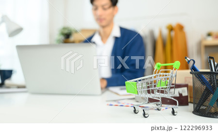 Small shopping cart on desk with entrepreneur working in background. Online retail and e-commerce concept Small shopping cart on desk with entrepreneur working in background. Online retail and e-commerce concept 122296933