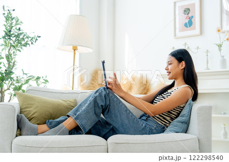 Side view and wide shot beautiful Asian woman lean on sofa also enjoy to use tablet in living room with day light. 122298540