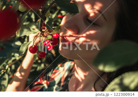 Close up portrait of a young woman plucking ripe red cherries from a tree in an orchard in summer. Harvesting, gardening, farming. Sun rays on a face. 122300914