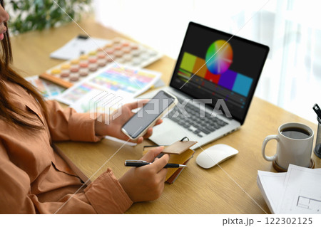 Female designer holding smartphone working with material and color samples at workplace 122302125
