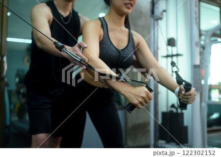 Young woman exercising on a cable machine in a modern gym with assistance from trainer 122302152