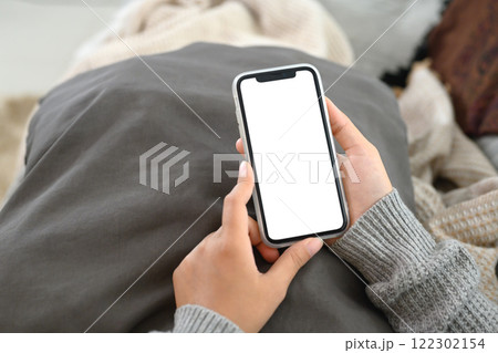 Hands holding smartphone with white screen while lounging under cozy blankets 122302154