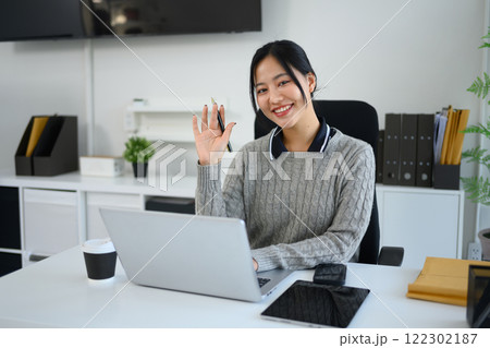 Smiling female employee sitting at her desk in a modern office and waving to the camera Smiling female employee sitting at her desk in a modern office and waving to the camera 122302187