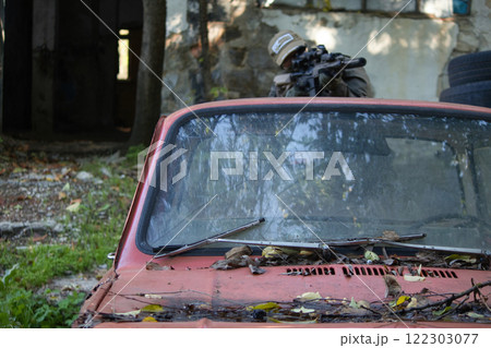 A tactical scene featuring a person in military gear aiming a rifle from behind cover provided by an old car.  122303077