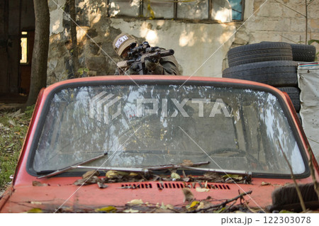 A tactical scene featuring a person in military gear aiming a rifle from behind cover provided by an old car. A tactical scene featuring a person in military gear aiming a rifle from behind cover provided by an old car. 122303078