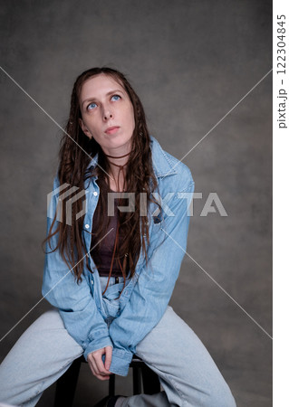 a beautiful and long-haired woman with big blue eyes in stylish clothes posing in a photo studio. the model sits on a chair and poses in denim clothes on a gray photo background. a beautiful and long-haired woman with big blue eyes in stylish clothes posing in a photo studio. the model sits on a chair and poses in denim clothes on a gray photo background. 122304845