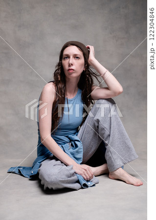 beautiful and long-haired woman in a hat and stylish clothes poses in a photo studio with a serious face. model sitting and posing in sportswear and barefoot on a gray photo background beautiful and long-haired woman in a hat and stylish clothes poses in a photo studio with a serious face. model sitting and posing in sportswear and barefoot on a gray photo background 122304846