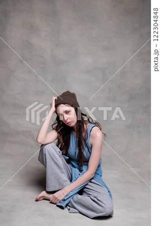 beautiful and long-haired woman in a hat and stylish clothes poses in a photo studio with a serious face. model sitting and posing in sportswear and barefoot on a gray photo background 122304848