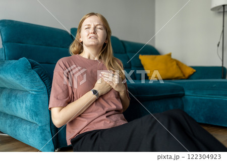 Woman sitting with strong chest pain and hand touching her chest while having trouble at home. Heart attack concept or heart failure symptom. Pressure in the chest. 122304923