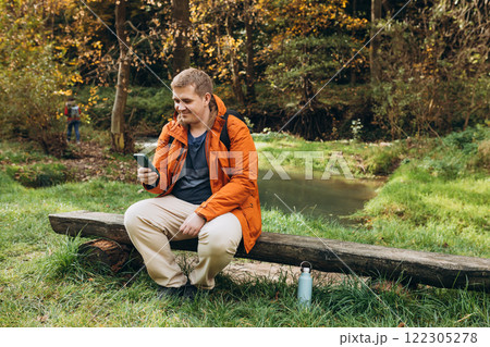 Stylish man with backpack holding smart phone and relaxing in nature. Full body photo. Hiking active male lifestyle siting on tree trunk outdoors. High quality photo. Adventures. 122305278