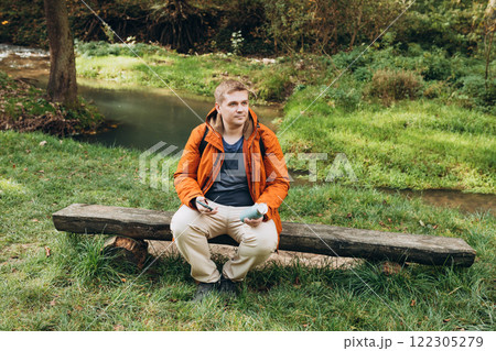 Stylish man with backpack holding smart phone and relaxing in nature. Full body photo. Hiking active male lifestyle siting on tree trunk outdoors. High quality photo. Adventures. 122305279