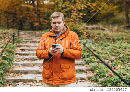 Adult 30s man hiker stand outdoors in a natural setting, smile while using his mobile phone outdoors. Hiking, lifestyle, adventure and tourism concept. Adult 30s man hiker stand outdoors in a natural setting, smile while using his mobile phone outdoors. Hiking, lifestyle, adventure and tourism concept. 122305427
