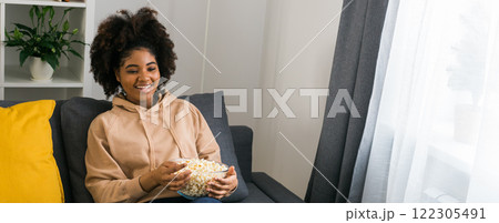Banner African American woman holds a bowl of popcorn while enjoying a television show. Bright smile radiates happiness and relaxation in a cozy home. TV, series and streaming service concept. Copy Banner African American woman holds a bowl of popcorn while enjoying a television show. Bright smile radiates happiness and relaxation in a cozy home. TV, series and streaming service concept. Copy 122305491