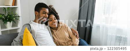 Banner copy space Happy African American couple enjoys quality time together while watching television. Man and woman seated closely on the couch, showcasing comfort and connection in a cozy home 122305495