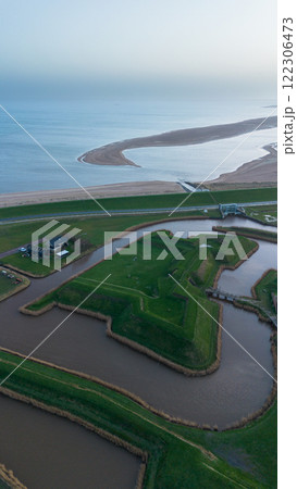 A drone video showing a unique landscape of man-made mounds and waterways near the sea, with a house on the grassy terrain, and a distant view of the coastline stretching under a cloudy sky. 122306473
