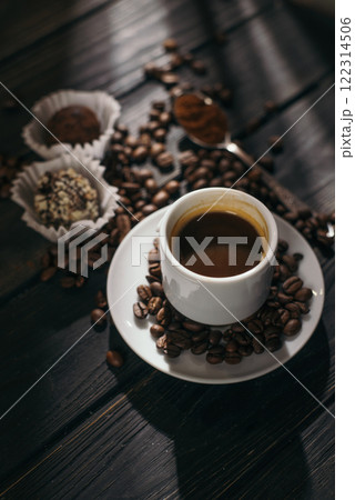 Cup of espresso surrounded by coffee beans on a dark wooden table, accompanied by chocolate truffles and a spoon with ground coffee. 122314506