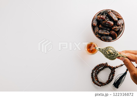 Hand pouring tea from a teapot with dates and prayer beads on white. Ramadan celebration 122316872