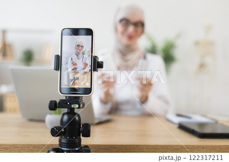 Middle Eastern Muslim woman recording video lesson on medicine. Engages audience through smartphone from home office setup with tripod, laptop, and tablet. 122317211