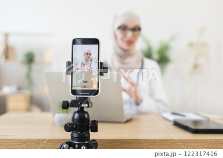 Young Muslim woman recording video lesson on phone for her blog. Laptop and tripod in modern workspace. 122317416