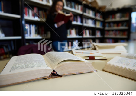 Stacks of open books lie on a table in a library as a woman reads in the background, symbolizing education, learning, and the pursuit of knowledge in an academic setting. Stacks of open books lie on a table in a library as a woman reads in the background, symbolizing education, learning, and the pursuit of knowledge in an academic setting. 122320344