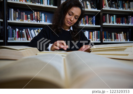 A focused young woman reads and studies among stacks of open books in a library. 122320353