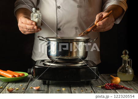 A skilled cook adds spices to a bubbling pot on a stovetop, balancing flavors with precision. Fresh vegetables and spices surround the warm setup, creating a homey cooking atmosphere A skilled cook adds spices to a bubbling pot on a stovetop, balancing flavors with precision. Fresh vegetables and spices surround the warm setup, creating a homey cooking atmosphere 122320554