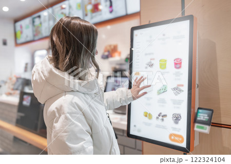 A 30 -35-year-old woman in a white winter jacket orders food through the screen menu at a fast food restaurant. A customer is utilizing a digital kiosk designed specifically for the purpose of 122324104
