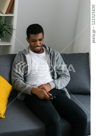 Cheerful curly haired ethnic african american male in casual clothes resting at home on comfortable sofa while watching tv channels or movie during weekend. Tv set, online television and entertiment 122325830