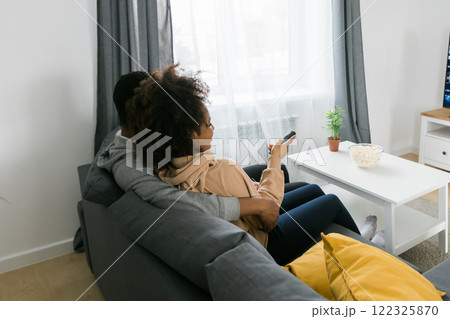 African American couple wife husband watching movie. Family pastime and leisure, watch television, rest. Relaxed man and woman watching TV series together sitting on couch at home enjoying eating African American couple wife husband watching movie. Family pastime and leisure, watch television, rest. Relaxed man and woman watching TV series together sitting on couch at home enjoying eating 122325870