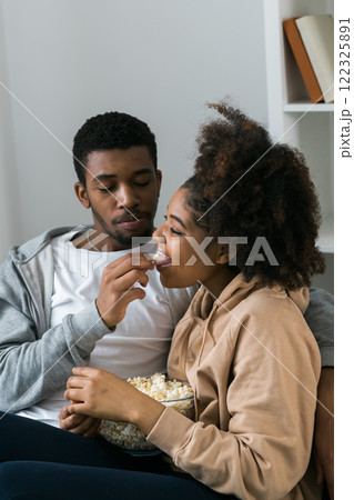 African American couple shares a playful and relaxed moment on the couch, with the man feeding popcorn to the woman. TV, series and streaming service concept 122325891