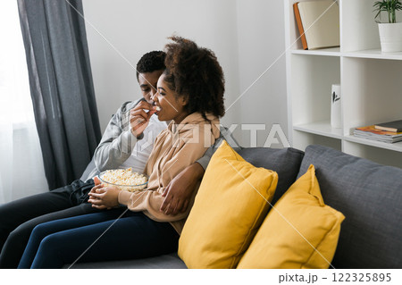 African American couple shares a playful and relaxed moment on the couch, with the man feeding popcorn to the woman. TV, series and streaming service concept 122325895