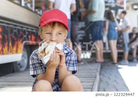Cute adorable pensive little boy kid enjoy eating hot dog sausage in bread near street cafe stall outdoors. Child healthy eating lunch hotdog. Junk food and fast food unhealthy snack concept 122329447
