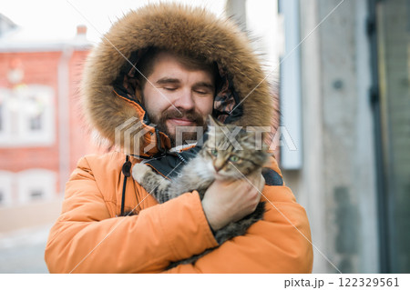 Bearded man in a fur lined hood smiles while holding a fluffy tabby cat outdoors. Cozy and joyful winter scene in an urban environment, homeless animals Bearded man in a fur lined hood smiles while holding a fluffy tabby cat outdoors. Cozy and joyful winter scene in an urban environment, homeless animals 122329561