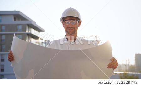 Male construction site manager, wearing safety helmets and glasses, is holding blueprints and inspecting a building site during sunrise or sunset, front side view. Architecture and engineering Male construction site manager, wearing safety helmets and glasses, is holding blueprints and inspecting a building site during sunrise or sunset, front side view. Architecture and engineering 122329745