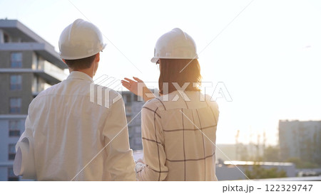 Two architects colleagues, wearing safety helmets and glasses, are examining blueprints at a construction site during sunrise or sunset, back view. Architecture and engineering Two architects colleagues, wearing safety helmets and glasses, are examining blueprints at a construction site during sunrise or sunset, back view. Architecture and engineering 122329747