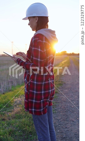 Woman engineer wearing a white protective helmet is taking notes with a clipboard in a field with wind turbines, as the sun sets. Clean energy and engineering concept Woman engineer wearing a white protective helmet is taking notes with a clipboard in a field with wind turbines, as the sun sets. Clean energy and engineering concept 122329775