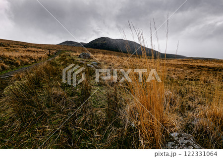 Grassy fields lead toward distant mountains under a dramatic sky filled with clouds. The landscape showcases earthy tones and rugged natural beauty. 122331064