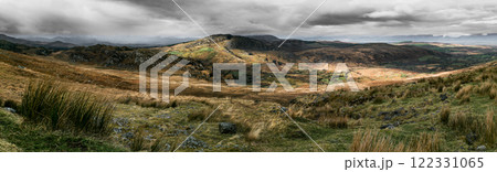 Rolling hills and mountains under a dark, brooding sky. Patches of green vegetation contrast with the brown landscape. A few small structures are visible. 122331065