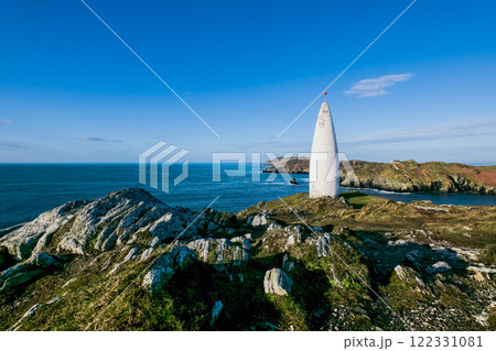 Coastal tower. A white, conical tower sits on a rocky, grassy headland, overlooking the sea. The day is bright and sunny. 122331081