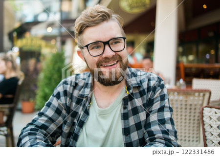 Bearded man in glasses laughing joyfully while sitting at a cafe table outdoors. 122331486
