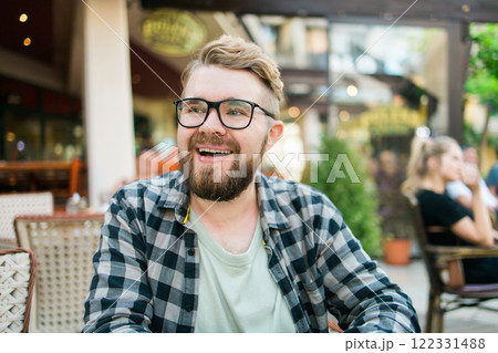 Bearded man in glasses laughing joyfully while sitting at a cafe table outdoors. Bearded man in glasses laughing joyfully while sitting at a cafe table outdoors. 122331488