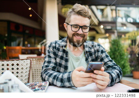 Smiling millennial man uses his smartphone while enjoying a casual outdoor cafe setting. Holidays atmosphere, modern connectivity, social networks 122331498