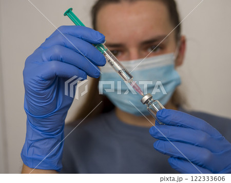 Healthcare professional preparing vaccine dose, wearing protective blue gloves and surgical mask, standing ready for immunization during coronavirus pandemic 122333606