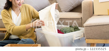 Smiling woman folding clothes for donation from a clear bin in her warm living room, promoting kindness through organization 122335325