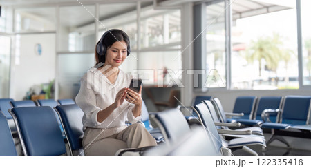 Happy young woman with headphones enjoying music while using smartphone in airport waiting area Happy young woman with headphones enjoying music while using smartphone in airport waiting area 122335418