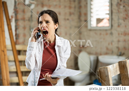 Irritated woman with papers talking on phone in house during renovation 122338118