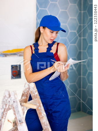 Portrait of young woman builder in blue construction overalls with documents in hands checks quality of completed repairs Portrait of young woman builder in blue construction overalls with documents in hands checks quality of completed repairs 122338443