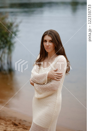 Caucasian woman in crochet beige dress standing on lake shore. Looking into camera. Caucasian woman in crochet beige dress standing on lake shore. Looking into camera. 122338733