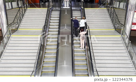 People ascending an escalator in a modern railway station. Bright, spacious public space with polished metal railings, organized infrastructure, and bustling transit movement. 122344376